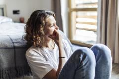 Une femme, assise sur le sol à côté de son lit, regarde dans le vide et semble soucieuse