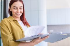 Une jeune femme aux cheveux roux sourit, tout en feuilletant un dossier au format papier