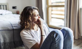 Une femme, assise sur le sol à côté de son lit, regarde dans le vide et semble soucieuse