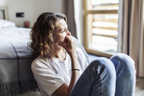 Une femme, assise sur le sol à côté de son lit, regarde dans le vide et semble soucieuse