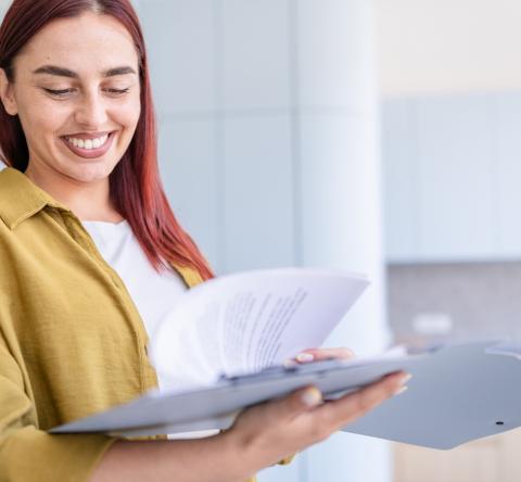Une jeune femme aux cheveux roux sourit, tout en feuilletant un dossier au format papier