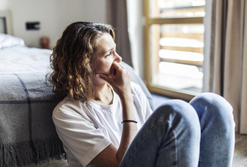 Une femme, assise sur le sol à côté de son lit, regarde dans le vide et semble soucieuse